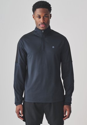Hombre vestido con un jersey negro de manga larga con cremallera hasta el pecho y pantalones cortos negros, de pie frente a un fondo gris claro liso.