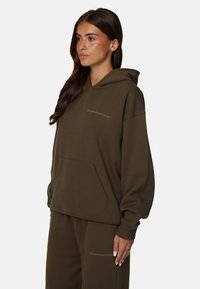 Oliwkowa bluza z kapturem, wykonana z miękkiego materiału, z kieszenią kangurką, luźny krój oraz haftowanym logo na klatce piersiowej. Zestawiona z pasującymi spodniami.