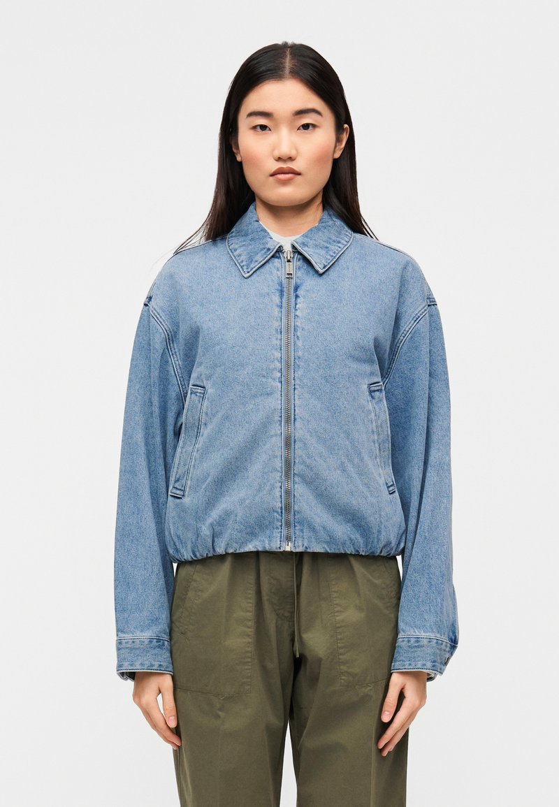 Ανοιχτό μπλε τζιν μπουφάν με γιακά, μπροστινό φερμουάρ και δύο πλαϊνές τσέπες. Χαλαρή εφαρμογή με ελαστικό τελείωμα και μακριά μανίκια.