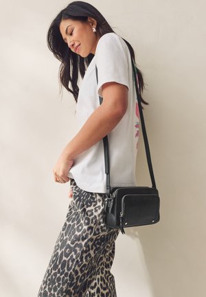 Crna kožna torbica preko tijela s dva pretinca na patentni zatvarač, nošena preko bijele majice i hlača s leopardim uzorkom, s jednostavnim dizajnom.