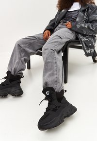 Zapatillas negras gruesas con puños forrados de piel, combinadas con jeans grises holgados. Los zapatos tienen una suela texturizada y detalles con costuras.