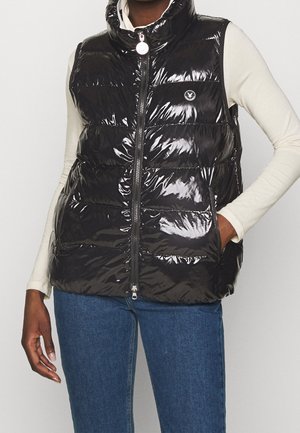 Personne portant une veste matelassée noire brillante avec fermeture éclair sur une chemise blanche à manches longues et un jean bleu, main dans la poche de la veste.