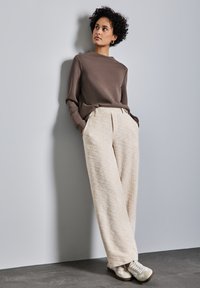 Brauner Pullover, locker geschnittene beige Designerhose und weiße Sneakers. Das Model lehnt an einer hellgrauen Wand und präsentiert das Outfit.