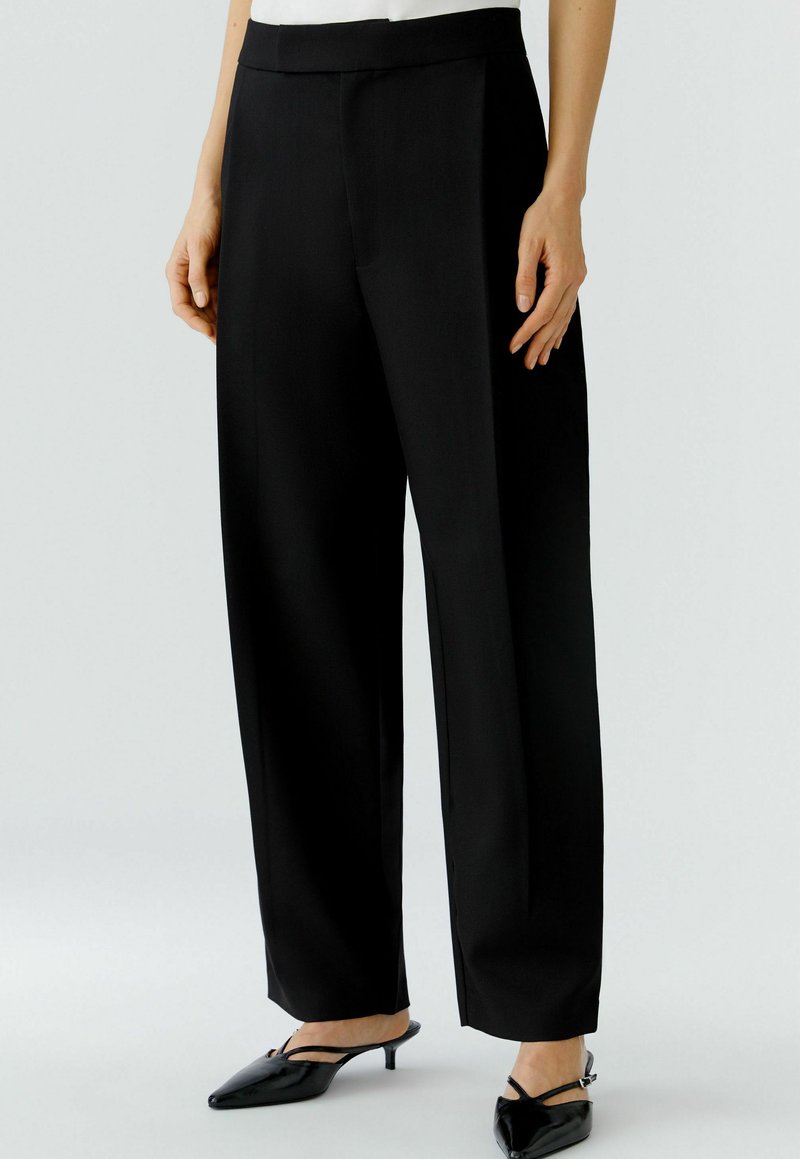 Zwarte wijde broek met een soepele textuur, voorzien van een hoge taille en voorplooien. Draagt met zwarte puntige hakken.