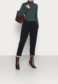 Person in einem grünen Rollkragenpullover, schwarzen ankle pants mit Umschlägen, braunen Ankle Boots aus Krokodil-Textur und mit einer passenden braunen Umhängetasche.