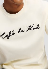 Cream-colored sweatshirt featuring embroidered black text "Café de Karl" across the chest, with a crew neck and soft, smooth texture.