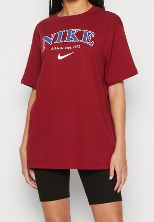 Kvinde iført en rød oversized Nike T-shirt med teksten "Athletic dept. 1972" og sorte ribbede cykelshorts, stående mod en ensfarvet baggrund.
