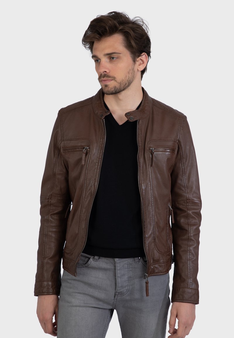 Brown leather jacket with a stand collar, zippered front, and two chest pockets. Features smooth texture and fitted design, paired with gray jeans.