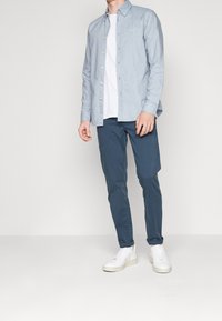 Světle modrá košile na knoflíky přes bílou tričko, v kombinaci s tmavě modrými kalhotami a bílými teniskami. Neformální outfit s uvolněným střihem.