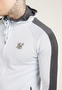Lys grå atletisk zip-up jakke med mesh tekstur, sorte detaljer på skuldrene og broderet logo på brystet. Hætte med justerbar snor.