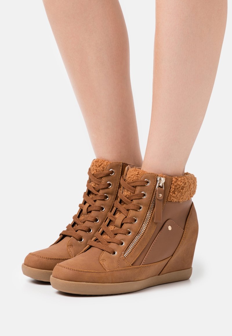 Bottines compensées marron à lacets sur le devant, fermeture éclair latérale et col en fausse fourrure, portées sur jambes nues sur fond blanc.