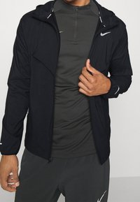 Mężczyzna w czarnej kurtce Nike noszący ciemnoszary rozpinany sportowy t-shirt i dopasowane spodnie, poprawiający suwak kurtki prawą ręką.