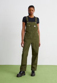 Olive green cotton overalls with adjustable straps, front pockets, and silver hardware, paired with a black T-shirt and chunky black boots.