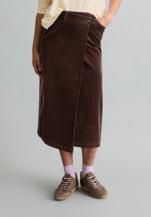 Persona che indossa una gonna midi in velluto a coste marrone con parte anteriore asimmetrica e sneakers marroni con calzini rosa su uno sfondo uniforme.