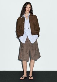 Brown suede cropped jacket over a light blue shirt, paired with brown suede knee-length shorts and black flip-flops on a plain background.