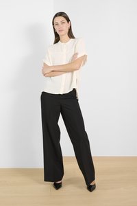 Sieviete stāv ar sakrustotām rokām, valkājot īsroku zīdkrekla krāsas kreklu ar pogām, melnas platajām kāju biksēm un melnas smailas kurpes uz koka grīdas.