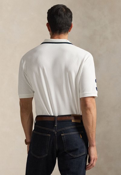 Άντρας που κοιτάζει προς τα πίσω, φοράει λευκό κοντομάνικο πόλο με σκούρα περιθώρια και σκούρο μπλε τζιν με καφέ δ�ερμάτινη ζώνη.