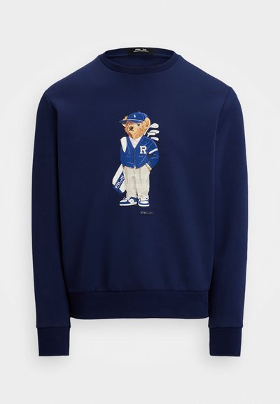 Sweatshirt bleu marine avec un graphisme d'un ours en tenue bleue et blanche avec des clubs de golf. Fabriqué à partir d'un tissu doux et texturé avec des poignets côtelés.