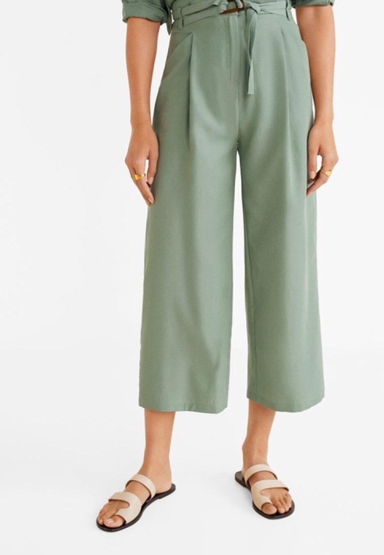 Femme portant un pantalon court taille haute, jambe large, vert sauge, avec une ceinture en tissu, associé à des sandales plates beiges à double bride.