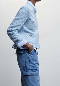 Veste en tweed bleu clair à double boutonnage avec boutons argentés. Associée à un pantalon cargo en jean bleu. Lignes épurées et tissu texturé.