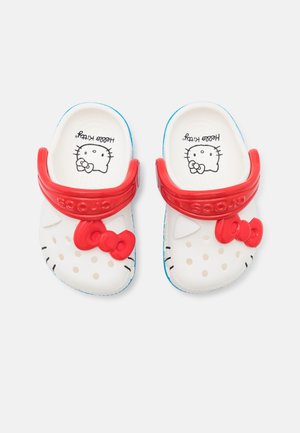 Witte Crocs met rode bandjes, voorzien van een verhoogde rode strik en Hello Kitty-ontwerp, versierd met kleine beige stippen en blauwe accenten.