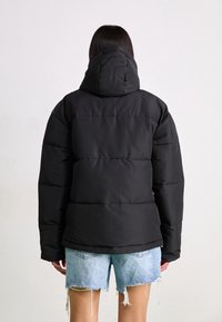 Persona con cabello largo y oscuro, vistiendo una chaqueta negra acolchada con capucha y shorts de mezclilla claros y deshilachados, de espaldas frente a un fondo sencillo.