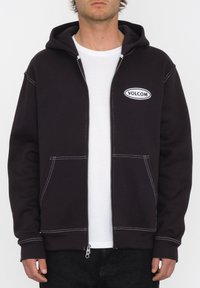 Haut zippé noir avec des coutures blanches, poches kangourou et un patch logo à l'avant. Dispose d'une capuche avec cordon de serrage, de poignets et d'un ourlet côtelés.