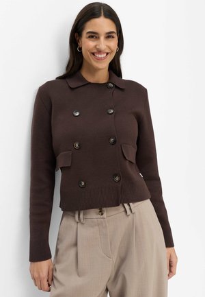 Junge Frau mit langen dunklen Haaren lächelt, trägt eine dunkelbraune, geknöpfte Jacke und beige hoch taillierte Hosen vor weißem Hintergrund.