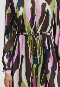 Mehrfarbiges Kleid mit einem braunen, grünen, pinken und weißen abstrakten Muster. Lange Ärmel, Knopfverschluss vorne, tailliert mit Zugband.