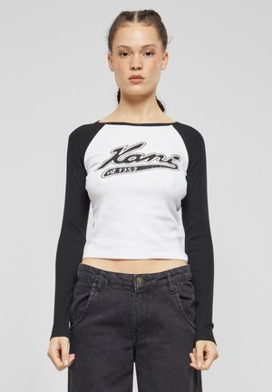 Hvit crop top med svarte raglanermer som har et front grafisk logo i svart. Brukt med høyt midje svarte jeans. Glatt tekstur.