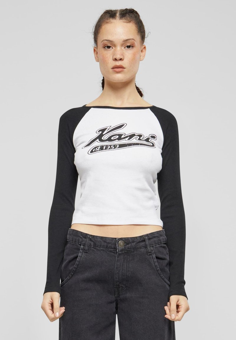 Bílý crop top s černými raglánovými rukávy, na přední straně s grafickým logem v černé barvě. Nosený s černými džínami s vysokým pasem. Hladká textura.