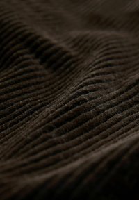 Close-up of soft, dark brown corduroy fabric showing raised, parallel ridges and fine texture in dim lighting.