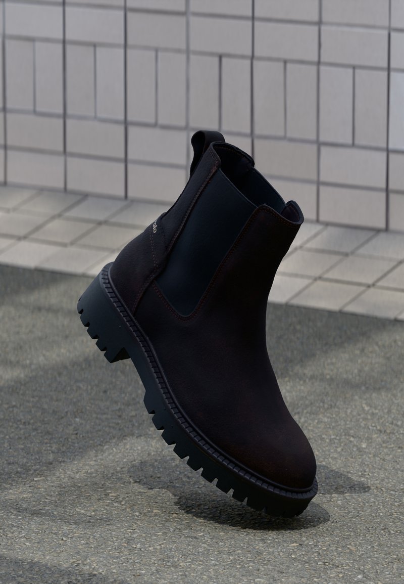 Bottes chelsea en cuir brun avec des panneaux élastiques noirs sur les côtés et une semelle épaisse et robuste. Couture remarquable autour des bords.