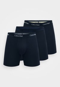Három pár sötétkék boxeralsó, puha anyagból készült, rugalmas derékrésszel, amelyen a "Calvin Klein" logó kontrasztos színekben szerepel.