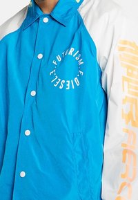 Modrá a bílá bunda se zapínáním na patentky, se textovými grafikami v oranžové a bílé barvě. Hladký povrch a raglánové rukávy.