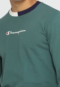 Felpa color verde teal realizzata in misto cotone, con scollatura rotonda rifinita con dettagli blu navy e avorio, e un piccolo logo ricamato sul petto.