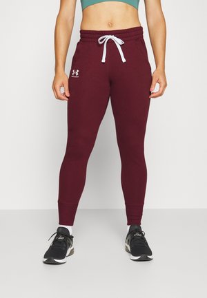 Person wearing maroon Under Armour joggers with white drawstring, black sneakers, and a teal cropped top, standing against a plain background.