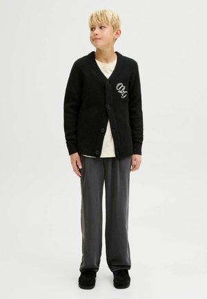 Black knitted cardigan with a V-neck, buttons, ribbed cuffs, and an embroidered logo. Paired with loose-fitting gray trousers and black slippers.