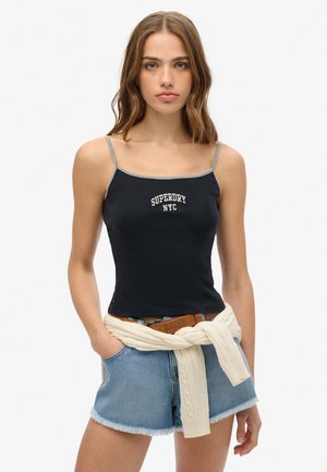 Débardeur noir côtelé avec bretelles fines, arborant le texte blanc "SUPERDRY NYC". Accompagné d'un short en jean bleu clair et d'un pull crème noué à la taille.