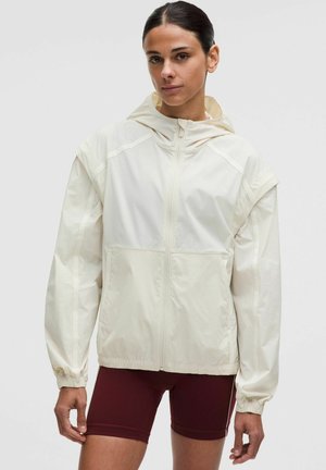 Vrouw draagt een lichtbeige capuchonvest met rits en elastische manchetten en donkerrode fietsshorts tegen een neutrale achtergrond.