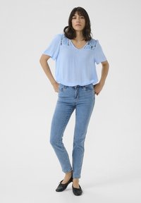 Blouse légère bleu clair à manches courtes avec des détails en dentelle au niveau du col, associée à un jean skinny à délavage moyen et des ballerines noires.