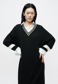 Črna pletena obleka z V-izrezom, z belimi in črnimi črtastimi poudarki na rokavih ter majhno podrobnostjo z logotipom.