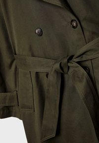 Manteau en daim vert foncé avec boutons croisés, ceinture nouée et coutures visibles sur les poches avant.