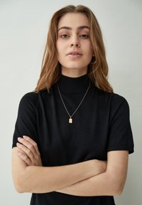 Černý pletený svetr s vysokým límečkem a krátkými rukávy; model nosí jemný zlatý přívěsek. Pozadí je neutrální a minimalistické.
