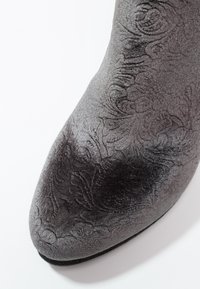 Gros plan d'un bout de chaussure en tissu gris avec des motifs floraux embossés complexes et un léger éclat sur fond blanc.