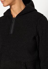Pullover in pile nero con cappuccio, caratterizzato da una mezza zip e un tessuto testurizzato. Il design è relaxed-fit con dettagli di tasca a marsupio.