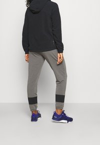 Felpa nera con cappuccio abbinata a pantaloni gris con polsini neri e scarpe da ginnastica blu, vista da dietro. Tessuto morbido, vestibilità casual.