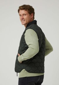 Chaleco acolchado verde oscuro con cuello negro, superpuesto sobre una camisa de manga larga verde claro, con bolsillos y dobladillo redondeado.