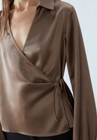 Brown satin blouse with a deep V-neck, wrap design, and long sleeves. Features adjustable side ties and smooth, glossy texture.