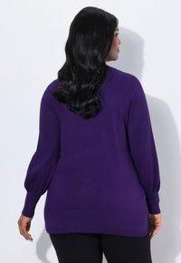 Maglione viola a maglia con maniche lunghe e svasate e orlo ondulato, caratterizzato da una texture liscia e una vestibilità slim. Indossato sopra pantaloni scuri.